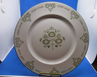 May include: A white ceramic dinner plate with a green floral design in the center and around the rim. The plate has a dark green border and is displayed on a gold-colored stand. The floral pattern includes stylized flowers and leaves.
