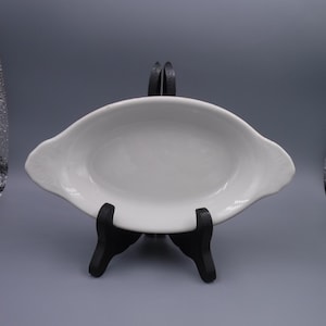 Może przedstawiać: Białe owalne naczynie do serwowania z uchwytami, prezentowane na czarnym stojaku. Naczynie ma gładką, błyszczącą powierzchnię i lekko podniesiony brzeg. Uchwyty są subtelnie zakrzywione.
