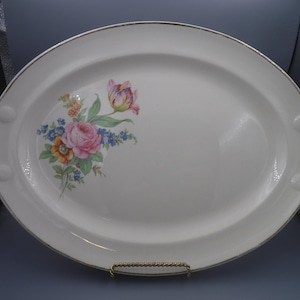 Puede incluir: Plato ovalado para servir con fondo blanco y diseño floral con rosas rosas, flores amarillas y naranjas y detalles azules. El plato tiene un borde dorado y asas.