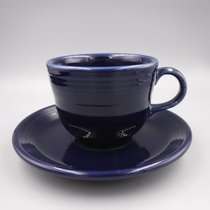 Puede incluir: Un juego de taza y platillo de cerámica azul marino. La taza tiene forma redondeada con asa, y el platillo es circular. Ambas piezas tienen un acabado brillante y están diseñadas con estrías horizontales.