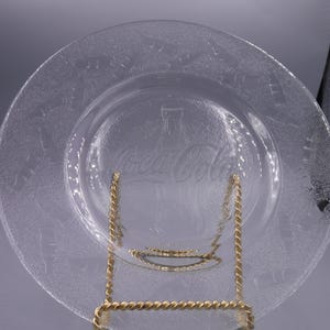 May include: Clear glass plate with Coca-Cola logo and bottle design. The plate is displayed on a gold-colored metal stand. The plate has a textured surface and a decorative border.