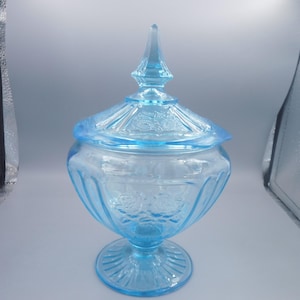 Puede incluir: Un cuenco de cristal azul claro con tapa a juego que presenta un remate puntiagudo. El cuenco y la tapa están decorados con patrones florales en relieve y nervaduras verticales, apoyados sobre una base circular con diseño de líneas.