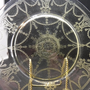 Puede incluir: Un plato de vidrio transparente con un diseño dorado. El diseño presenta un patrón repetitivo de cadenas y motivos florales.