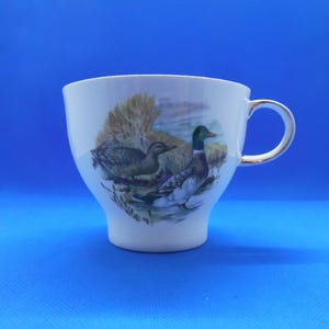 May include: White ceramic teacup with a gold rim and a design of two ducks on a lake. The ducks are green, brown, and white.