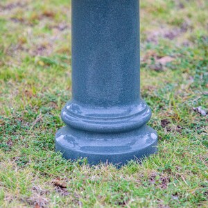 May include: A dark teal, decorative pillar with a textured surface. The pillar has a rounded base and a cylindrical body. It is set in a bed of green grass.
