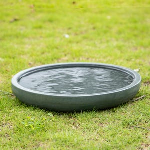 May include: A round, gray bird bath filled with water, set on a bed of green grass. The bird bath has a simple, modern design with a wide, shallow basin. The water's surface reflects the sky.