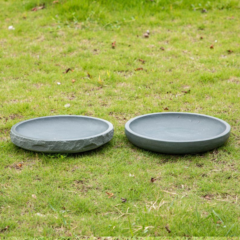 以下が含まれることがあります： 緑の草の上に置かれた、2つの灰色の石のようなバードバス。 バードバスは丸く浅く、粗い質感の縁があります。 鳥に水を与えるように設計されています。