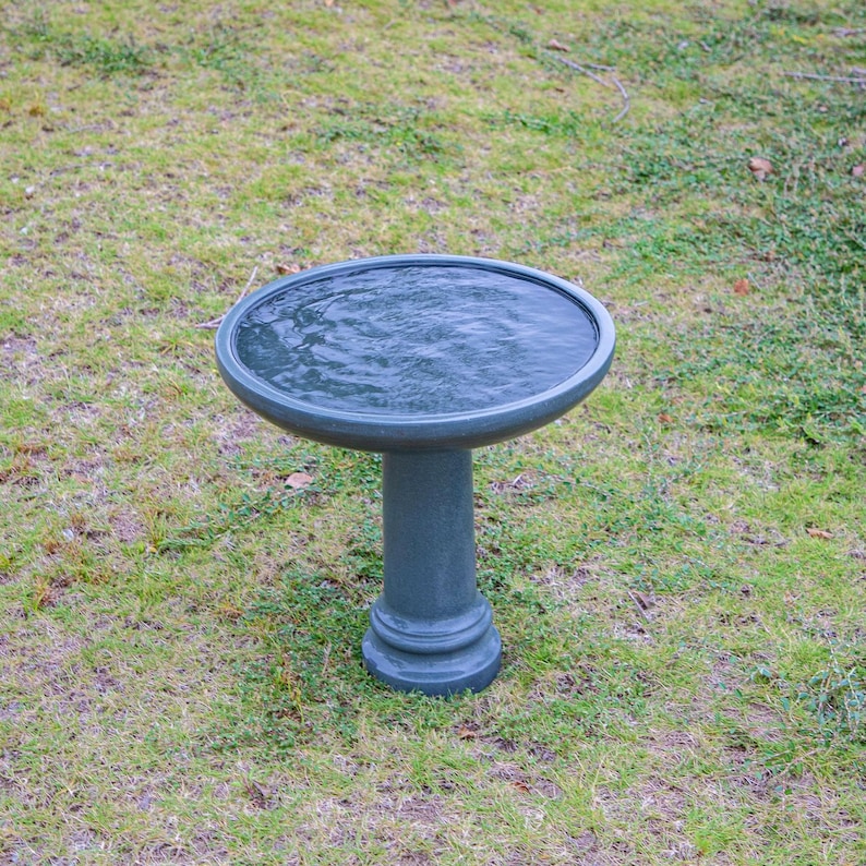 May include: A dark teal bird bath with a round basin filled with water. The bird bath has a pedestal base and is set on a grassy lawn. The water's surface shows slight ripples, suggesting recent activity or wind.