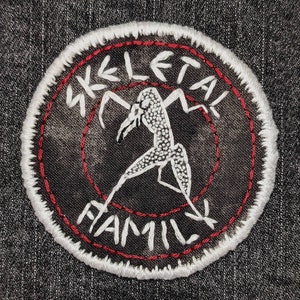 Puede incluir: Parche blanco bordado con costuras rojas. El parche presenta una figura de esqueleto con las palabras "SKELETAL FAMILY" alrededor.