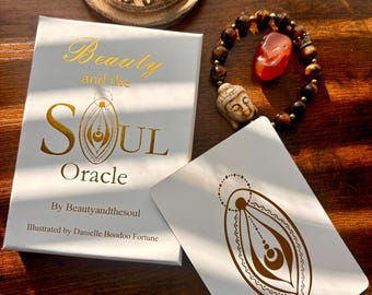 Op de afbeelding: Een Beauty and the Soul Oracle kaartendeck en een enkele kaart met goudfolie ontwerpen, een kralenarmband met een uitgesneden Boeddha-hoofd en een rode steen, en een ronde houten wierookbrander met een rokende wierookkegel op donker hout.