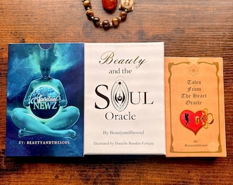 May include: An arrangement featuring three oracle card decks and a beaded bracelet with a Buddha head charm. The decks are titled "Spiritual Newz", "Beauty and the Soul Oracle", and "Tales From The Heart Oracle". The bracelet includes a red stone.