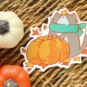May include: A sticker featuring a cartoon cat wearing a teal scarf and a crown, hugging three orange pumpkins. The sticker is surrounded by fall leaves. Decorative pumpkins in orange and white are also visible.