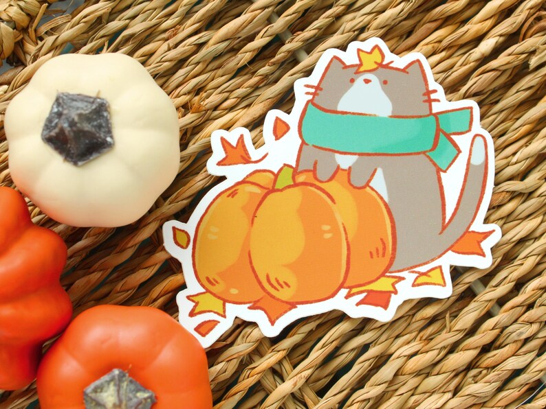 May include: A sticker featuring a cartoon cat wearing a teal scarf and a crown, hugging three orange pumpkins. The sticker is surrounded by fall leaves. Decorative pumpkins in orange and white are also visible.