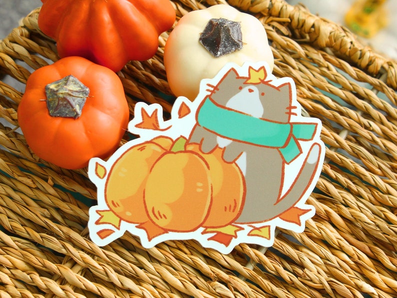 May include: A sticker featuring a cartoon cat wearing a teal scarf and a crown, hugging three orange pumpkins. The sticker has a white border and is decorated with fall leaves. Decorative pumpkins are in the background.