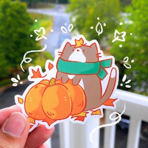 May include: A sticker featuring a cartoon cat wearing a green scarf and a crown, perched atop two orange pumpkins. The cat is brown and white, surrounded by autumn leaves. Whimsical white doodles accent the design.