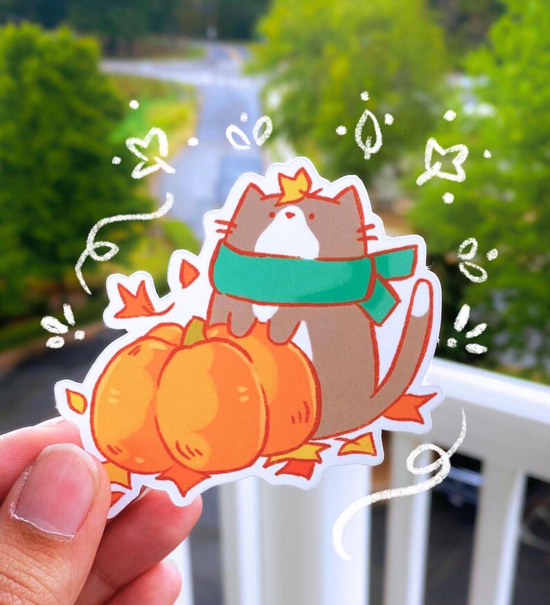 May include: A sticker featuring a cartoon cat wearing a green scarf and a crown, perched atop two orange pumpkins. The cat is brown and white, surrounded by autumn leaves. Whimsical white doodles accent the design.
