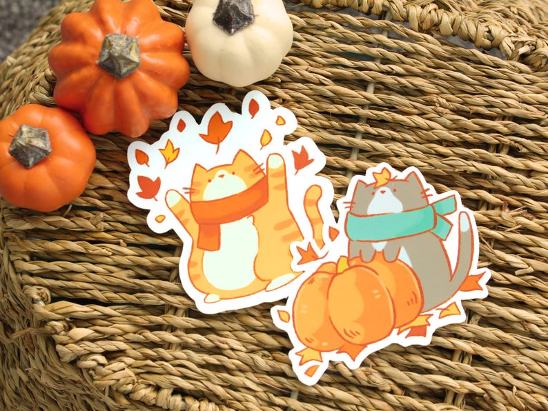 May include: Two cat stickers with autumn themes, one orange and one gray, wearing scarves and surrounded by leaves. Small pumpkins in orange and white are arranged around the stickers on a woven surface.