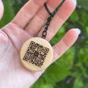May include: A wooden keyring with a QR code and the word "Hi" engraved on it. The keyring has a black metal ring and chain. The round wooden piece is light brown. The background is blurred green foliage.