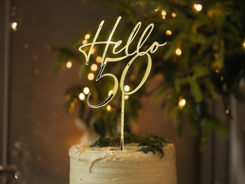 Gold cake topper with the text "Hello 50" on a white frosted cake with green foliage.