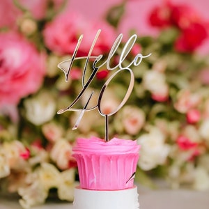 May include: A pink cupcake with white frosting and a gold topper that says "Hello 40" in front of a pink and white floral background.