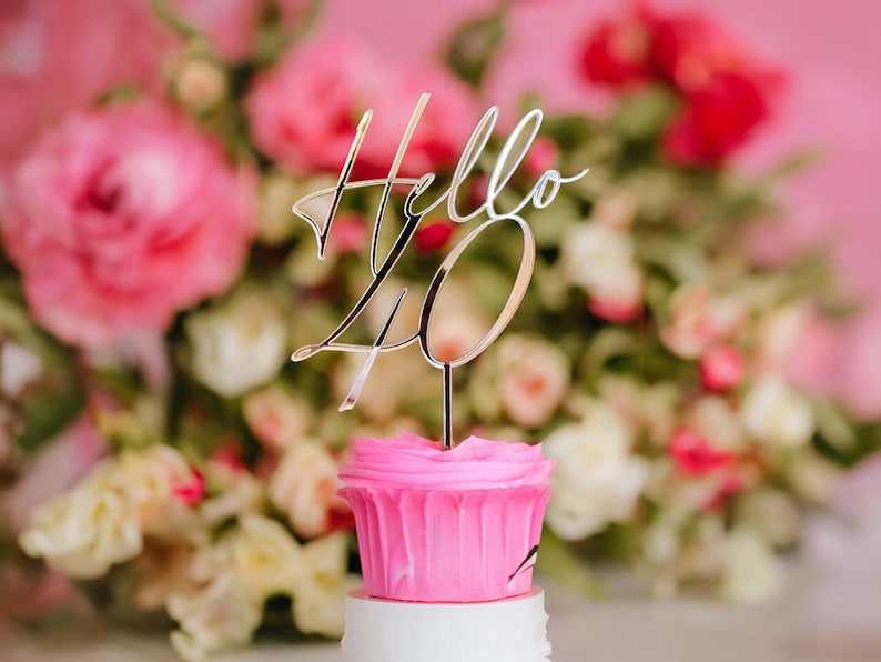 May include: A pink cupcake with white frosting and a gold topper that says "Hello 40" in front of a pink and white floral background.
