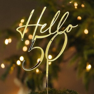 May include: Gold cake topper with the words "Hello 50" in a cursive font. The topper is placed on a white frosted cake with green foliage.