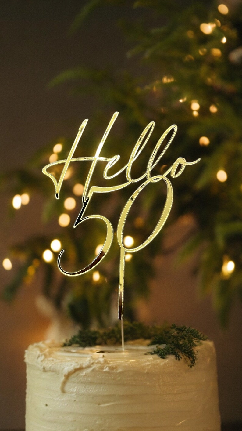 May include: Gold cake topper with the words "Hello 50" in a cursive font. The topper is placed on a white frosted cake with green foliage.