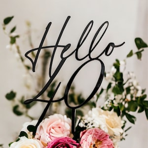 May include: A white cake with pink and white flowers and a black acrylic cake topper that says "Hello 40".