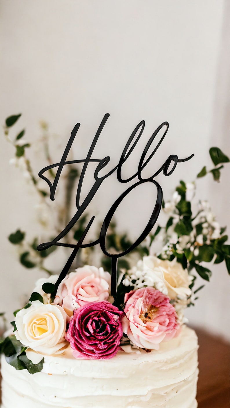 May include: A white cake with pink and white flowers and a black acrylic cake topper that says "Hello 40".