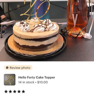 May include: Gold cake topper with the words "Hello Forty" on it. The topper is on top of a layered cake with chocolate and white frosting. A bottle of pink wine is in the background.