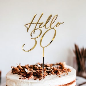 May include: Gold cake topper with the words "Hello 30" in a cursive font. The topper is on a cake with brown frosting and chocolate sprinkles.