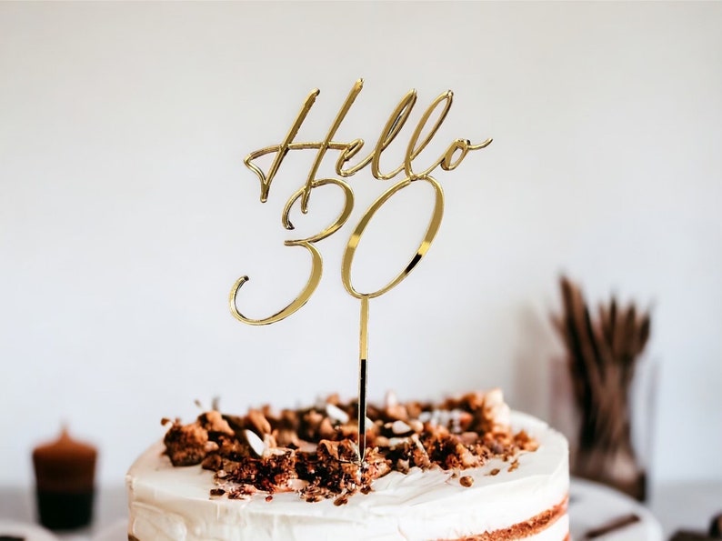 May include: Gold cake topper with the words "Hello 30" in a cursive font. The topper is on a cake with brown frosting and chocolate sprinkles.