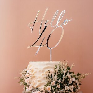 May include: A white cake with a silver topper that says "Hello 40" and is decorated with white flowers.