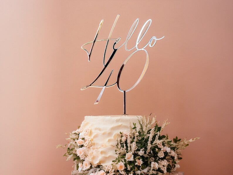 May include: A white cake with a silver topper that says "Hello 40" and is decorated with white flowers.