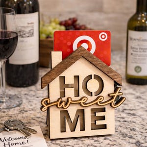 Può includere: Portacarte a forma di casa in legno con le parole "HOME sweet ME" ritagliate. Una carta regalo rossa è inserita nel supporto. Bottiglie di vino e un bicchiere di vino rosso sullo sfondo.