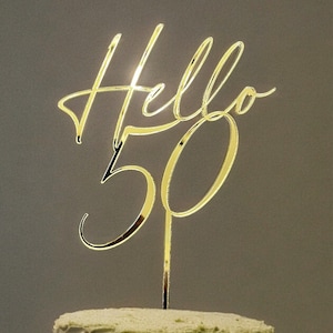 May include: Gold cake topper with the words "Hello 50" in a cursive font. The topper is on top of a green frosted cake.