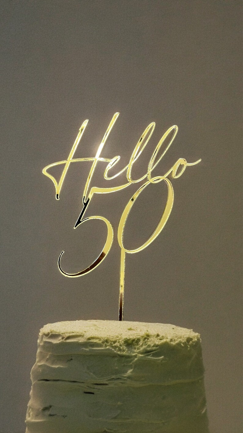 May include: Gold cake topper with the words "Hello 50" in a cursive font. The topper is on top of a green frosted cake.