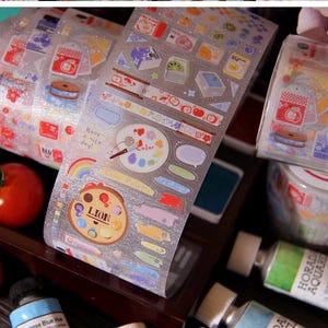 May include: Rolls of decorative washi tape with colorful illustrations, including food, art supplies, and speech bubbles. The tape features a clear, glittery background. The image also includes fresh tomatoes and art supplies.