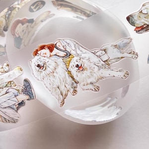May include: Clear sticker roll featuring illustrations of a person with red hair and two fluffy white dogs. The dogs have their tongues out. Other illustrations of dogs and people are visible on the roll.