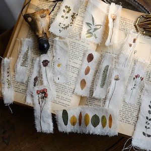 May include: A collection of vintage-style fabric scraps with pressed botanicals and text printed on them. The scraps feature pressed leaves, flowers, and ferns in shades of green, brown, and yellow. The text is in French and includes words like "extra" and "vaguant".