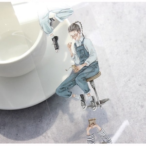 May include: Watercolor illustration of a person in denim overalls, seated on a stool, holding a small object. Another figure is partially visible above. The image has a soft, artistic style, with a neutral background and a white teacup.