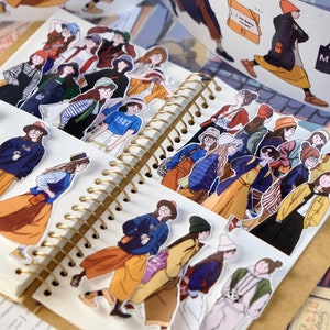May include: A collection of stickers featuring illustrations of women in various outfits, including coats, hats, and skirts. The stickers are arranged on an open spiral notebook with a gold binding. The color palette includes shades of blue, orange, and yellow.