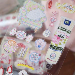 May include: Clear sticker sheet with various illustrations, including a floral armchair, candies, a coffee cup, and a tube of English Rose cream. The tube is labeled "English Rose" and contains 75ml of product. Other text includes "Rose" and "about you,".