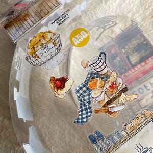 May include: A clear sticker sheet with watercolor illustrations of baked goods and coffee. Includes a basket of bread, a pastry with a cherry, a checkered pitcher, and a coffee cup. A yellow circle with the number "A50" is also visible.