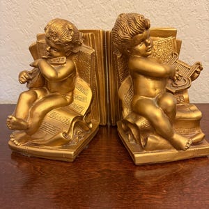 Puede incluir: Sujetalibros dorados con figuras de querubines tocando instrumentos musicales. Cada querubín está sentado y sostiene un instrumento de cuerda, con notas musicales visibles en los sujetalibros. Los sujetalibros son de metal.