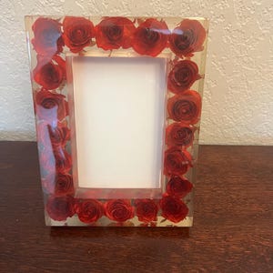 Może przedstawiać: Prostokątna ramka na zdjęcia z przezroczystą żywiczną obwódką, z zatopionymi czerwonymi różami. Ramka ma białe wnętrze do wyświetlania zdjęcia i jest umieszczona na drewnianej powierzchni.