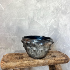Puede incluir: Un cuenco de cerámica hecho a mano con un exterior oscuro y texturizado y un interior azul oscuro. El cuenco tiene una forma única y orgánica con detalles en relieve en forma de botón. Se encuentra sobre un taburete de madera rústica y envejecida.