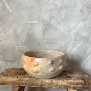 May include: A white ceramic bowl with a textured, bumpy surface. The bowl has a light orange and brown glaze and is sitting on a rustic wooden stool.
