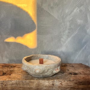 Könnte beinhalten: Eine kleine, weiße Kerze in einem grauen, strukturierten Keramikbehälter mit einem Holzdocht. Die Kerze steht auf einer rustikalen Holzoberfläche.