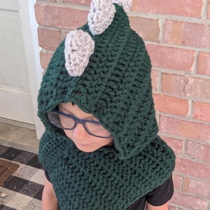 May include: A dark green crocheted hooded cowl featuring three light gray crocheted spikes along the top ridge. The hood is constructed with a thick yarn texture and covers the head, extending down into a pointed triangular shape over the chest.